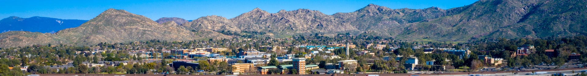 A wide panoramic view of a the UC Riverside campus nestled at the base of rugged, rocky mountains under a clear blue sky. The landscape features a mix of academic buildings, green spaces, and palm trees stretching across the valley floor.