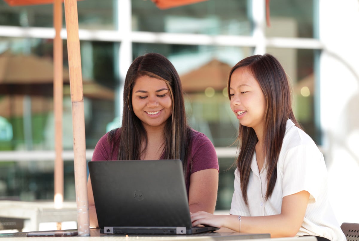 Two smiling female students work together on a laptop at a sunny outdoor table on the UCR campus.