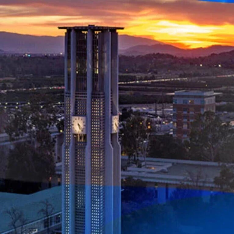 Aerial view of the Bell Tower on the UCR campus. The sun is setting behind the mountains in the background giving off a bright orange light.