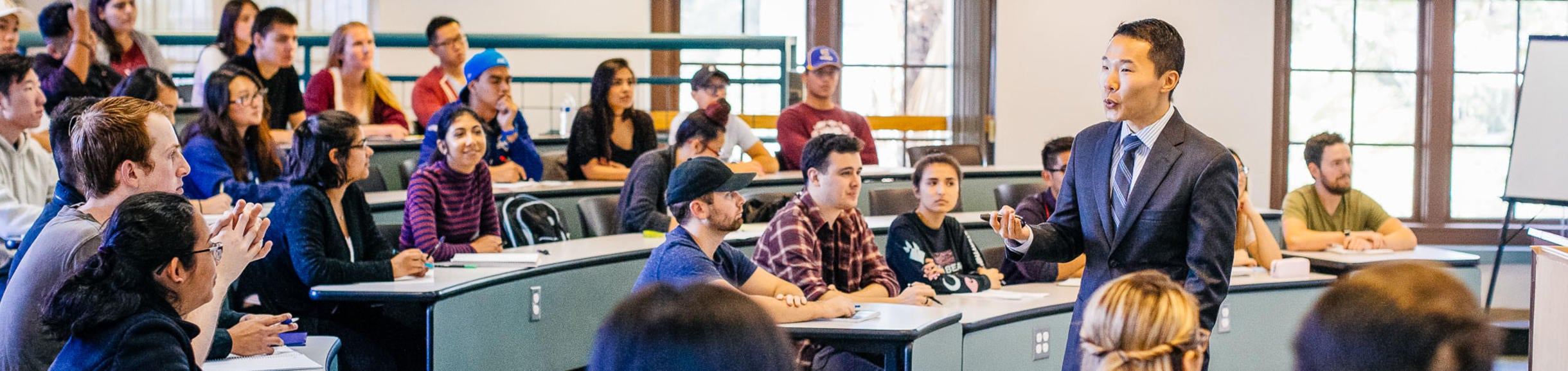 A classroom of students sitting at desks listen to a lecture by a man in a suit.