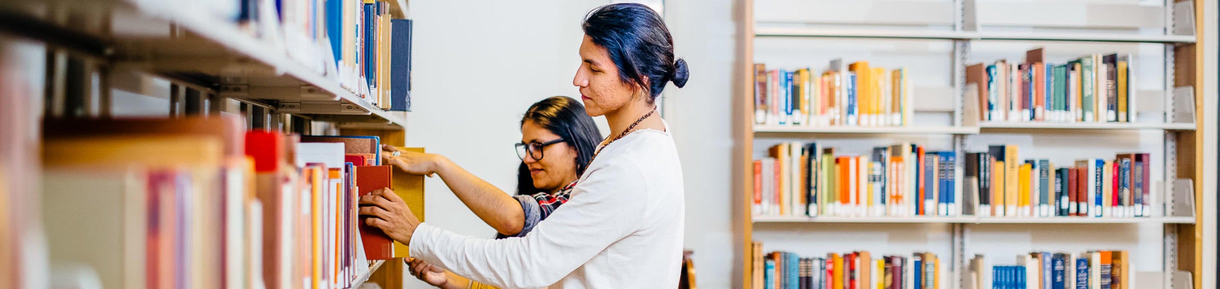 A man and a woman look at books that sit on a shelf in a library. Other shelves of books are around them.